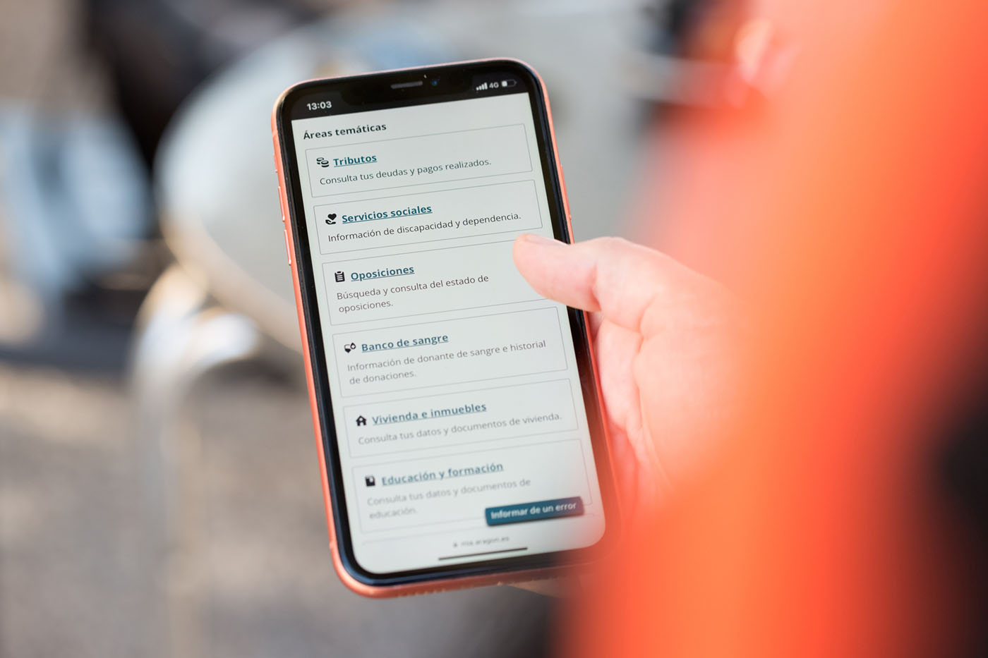 Una persona sostiene en la mano un teléfono móvil. En la pantalla se ven las áreas temáticas de MiA, que son las secciones de la app organizadas por temas. La primera de las áreas es "Tributos".