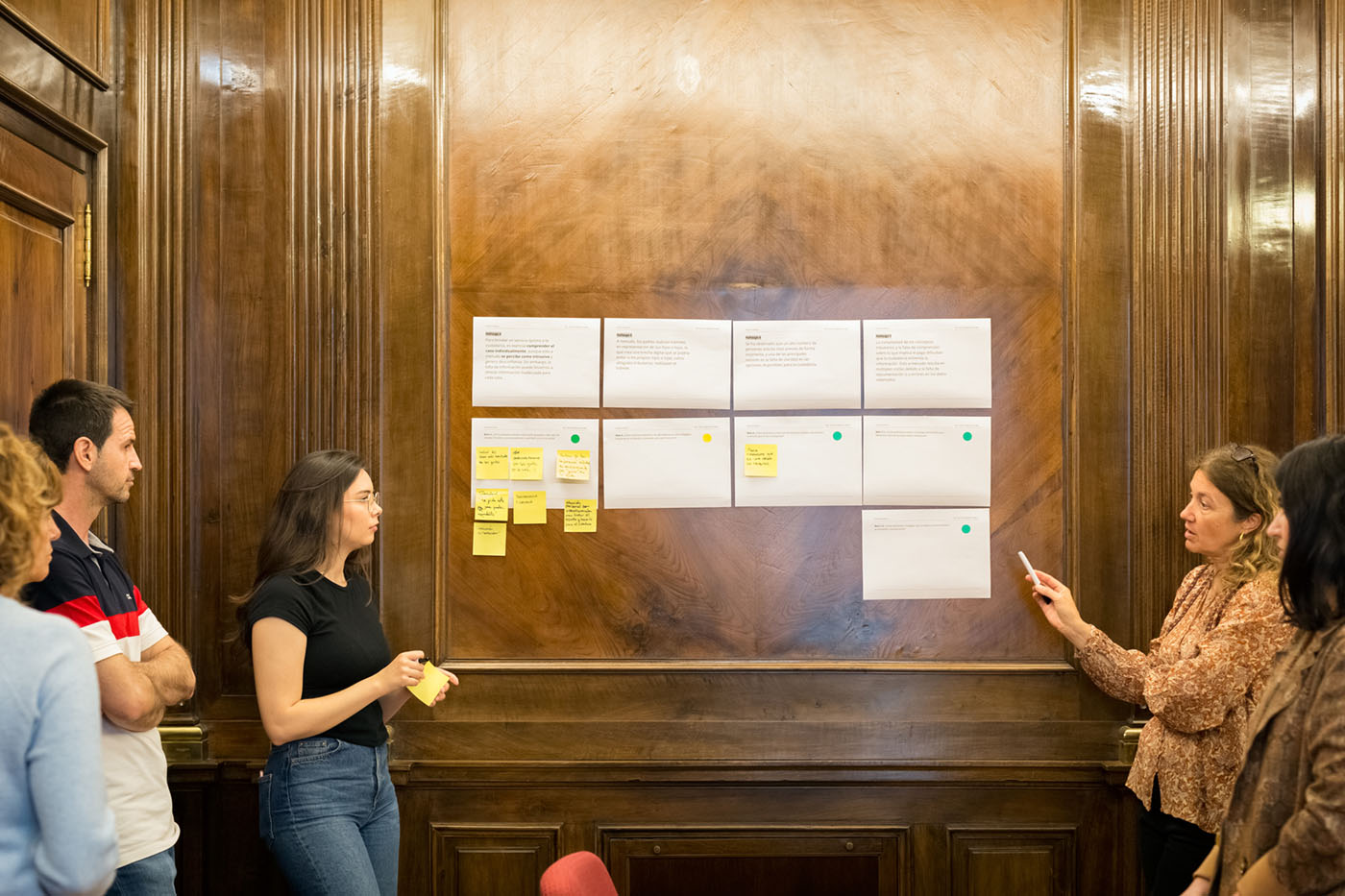 Varias personas están reunidas al rededor de unos paneles pegados en la pared, algunos con notas adhesivas pegadas. La persona más visible a la izquierda lleva en la mano un taco de notas adhesivas y está en actitud de escuchar. La persona más visible a la derecha señala y está en actitud de hablar.