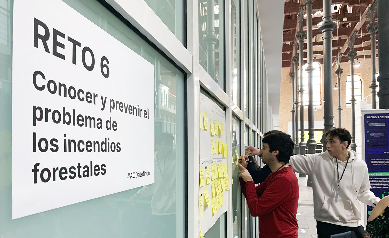 En primer plano se lee la descripción de uno de los retos, en segundo plano dos hombres jóvenes están escribiendo sus ideas en post-its y pegándolas en la pared