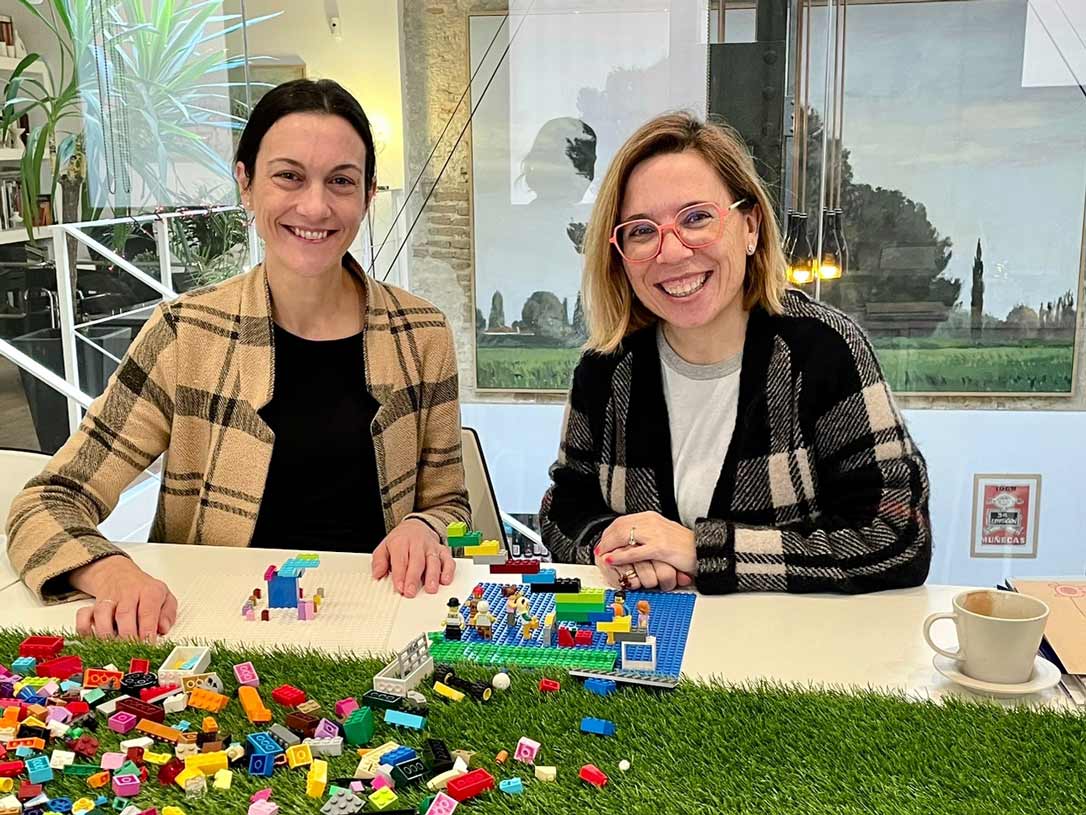 Dos mujeres con piezas de Lego en la mesa y sonriendo a cámara